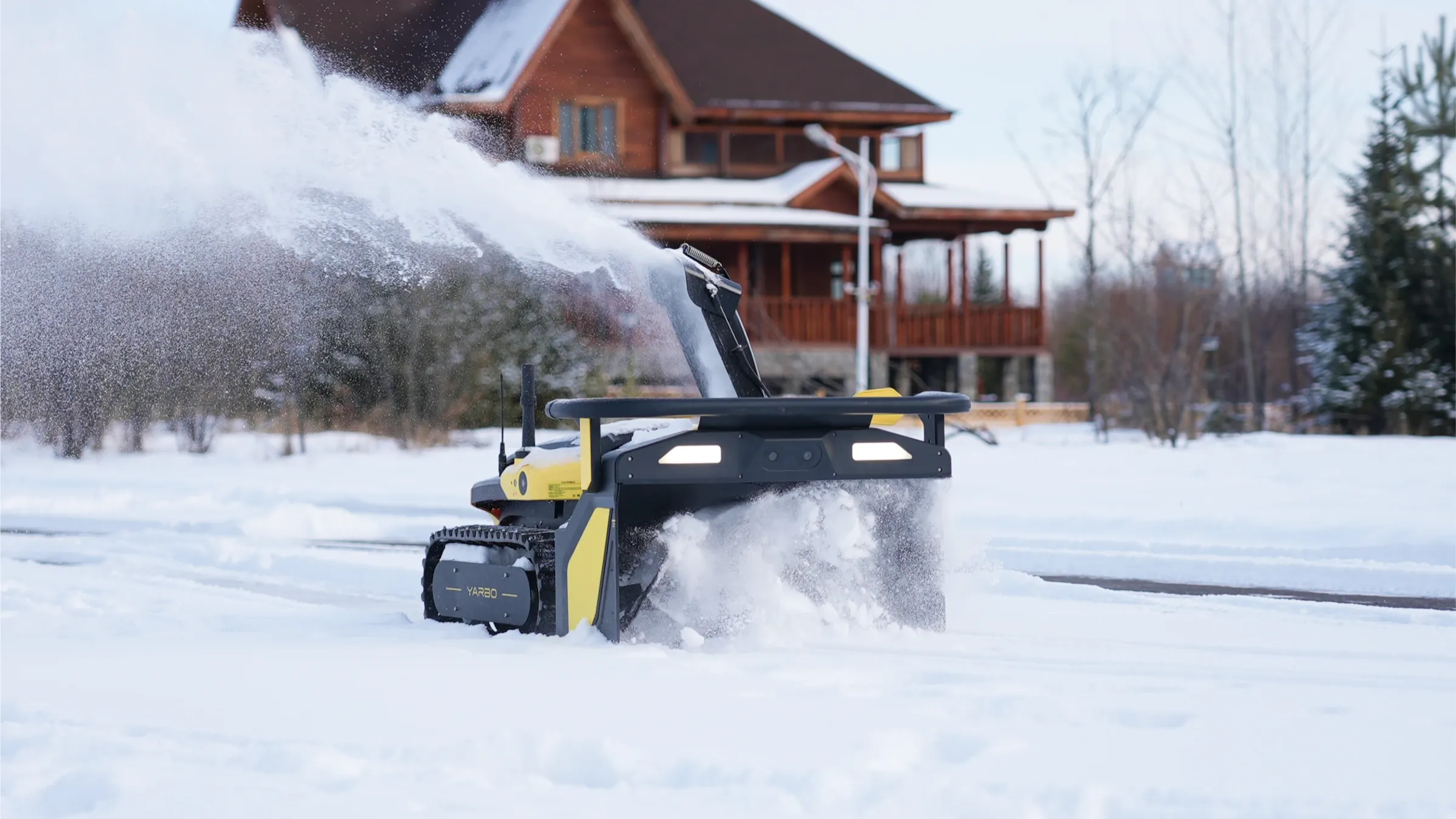 Robot Yarbo avec module souffleuse projetant la neige devant une maison en hiver
